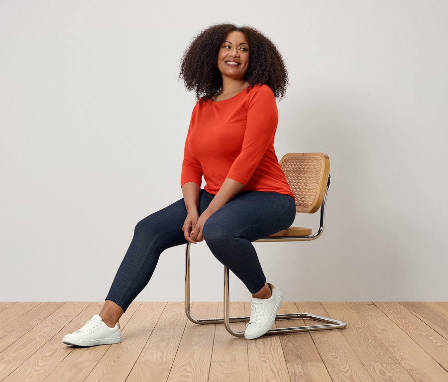 Une femme est assise sur une chaise, portant un chemisier orange, un legging bleu marine et des baskets blanches.