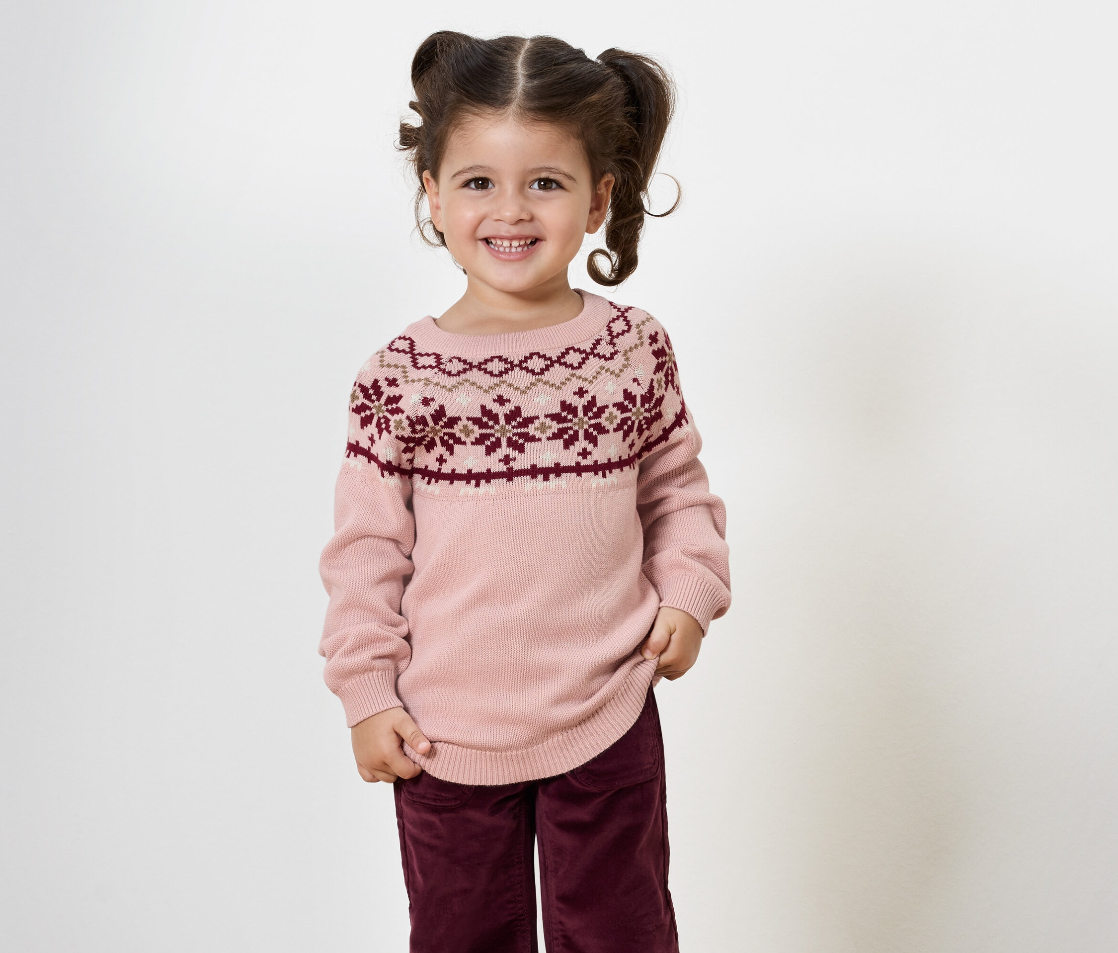 Petite fille souriante avec des couettes posant dans un pull rose à motifs et un pantalon bordeaux.
