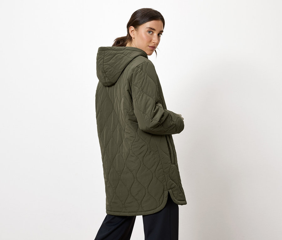 Une femme regarde par-dessus son épaule et porte un manteau matelassé vert olive à capuche.