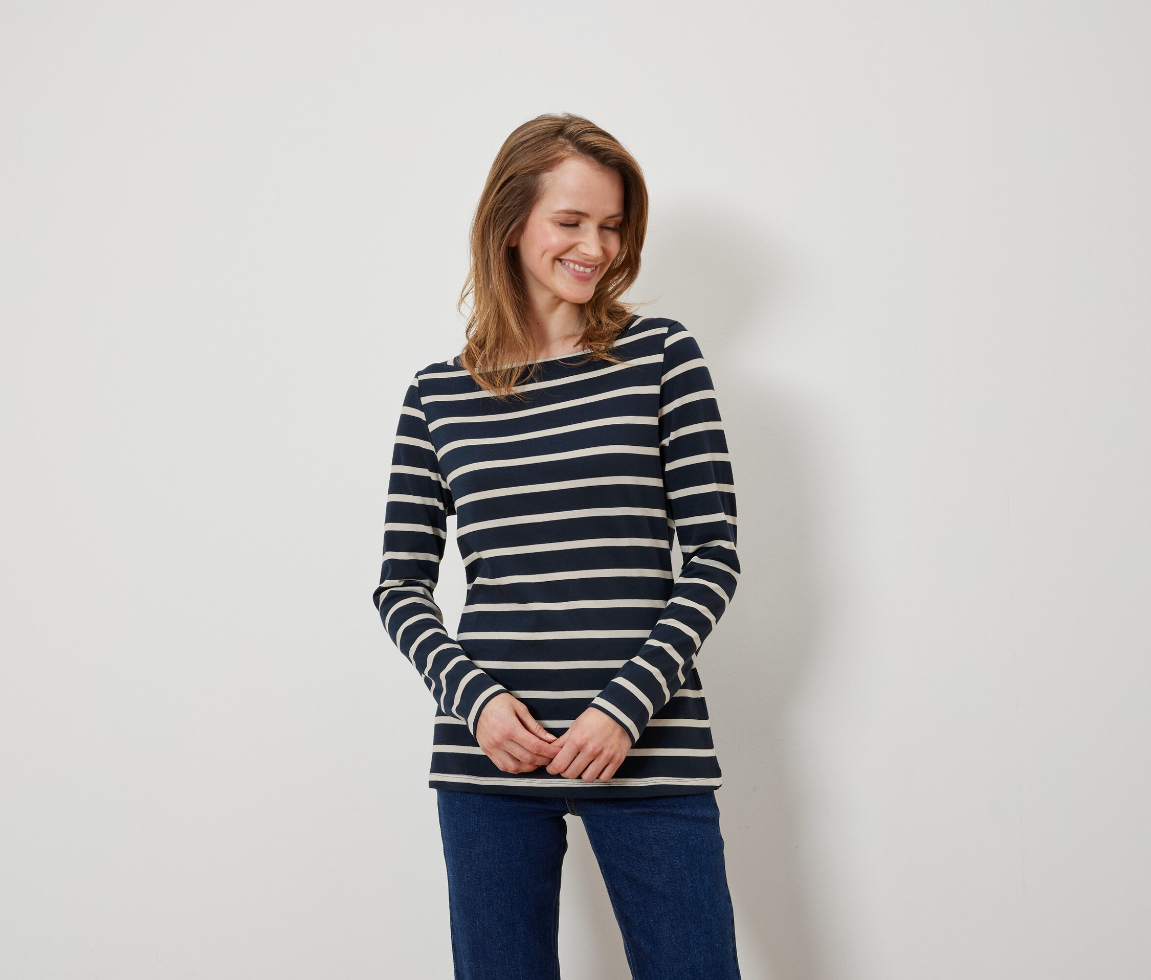 Une femme aux cheveux bruns et aux yeux bleus porte un haut à manches longues rayé bleu et blanc et un jean bleu.