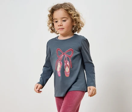 Fillette aux cheveux bouclés portant un T-shirt gris avec un motif de chaussons de ballet et un pantalon rose.