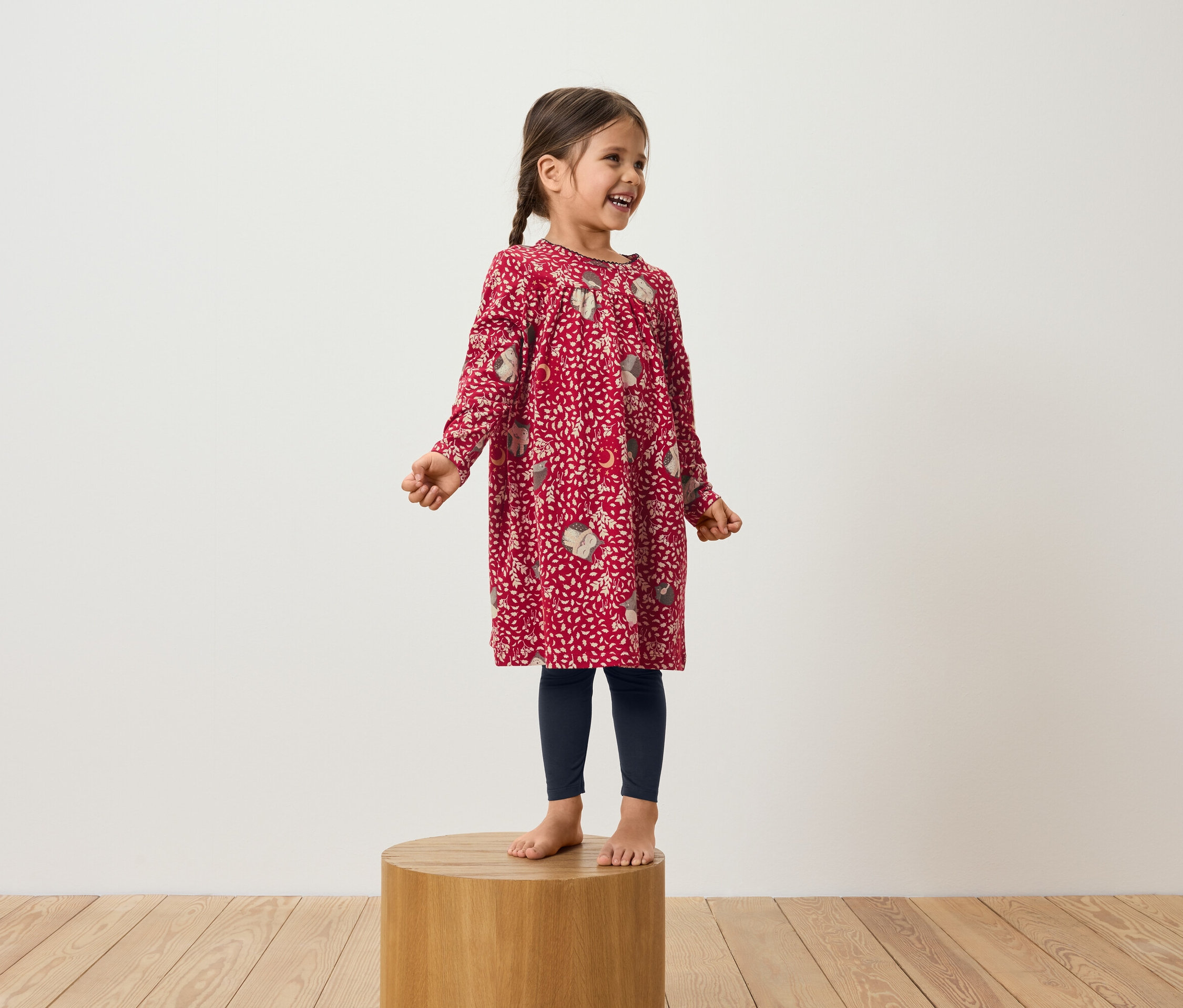 Une fillette souriante se tient sur un socle en bois, portant une robe rouge à manches longues et des leggings bleu marine.