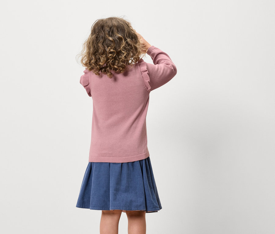 Une fille aux cheveux bouclés se tient dos à l'objectif, portant un pull rose et une jupe bleue.