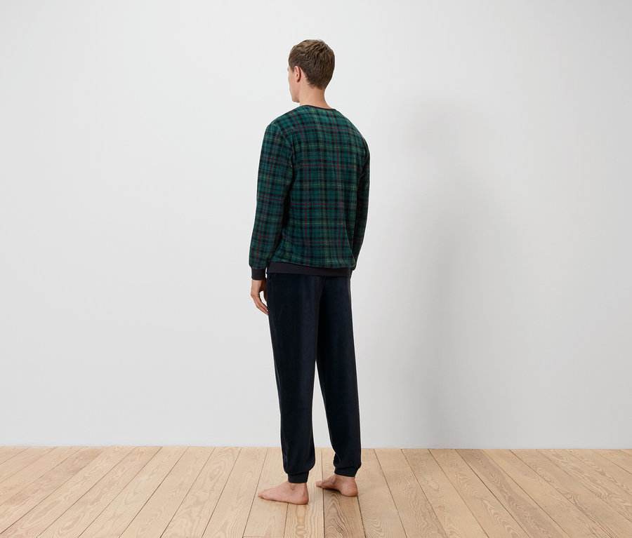 Un homme, dos à la caméra, porte un haut à carreaux vert et noir et un pantalon bleu marine, debout sur un plancher en bois devant un mur blanc.