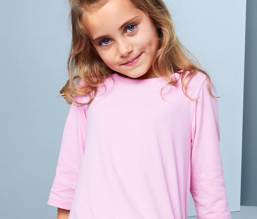 Une fille aux cheveux bruns et aux yeux bleus porte une robe rose. Elle a 3 élastiques « chouchou » dans les cheveux.