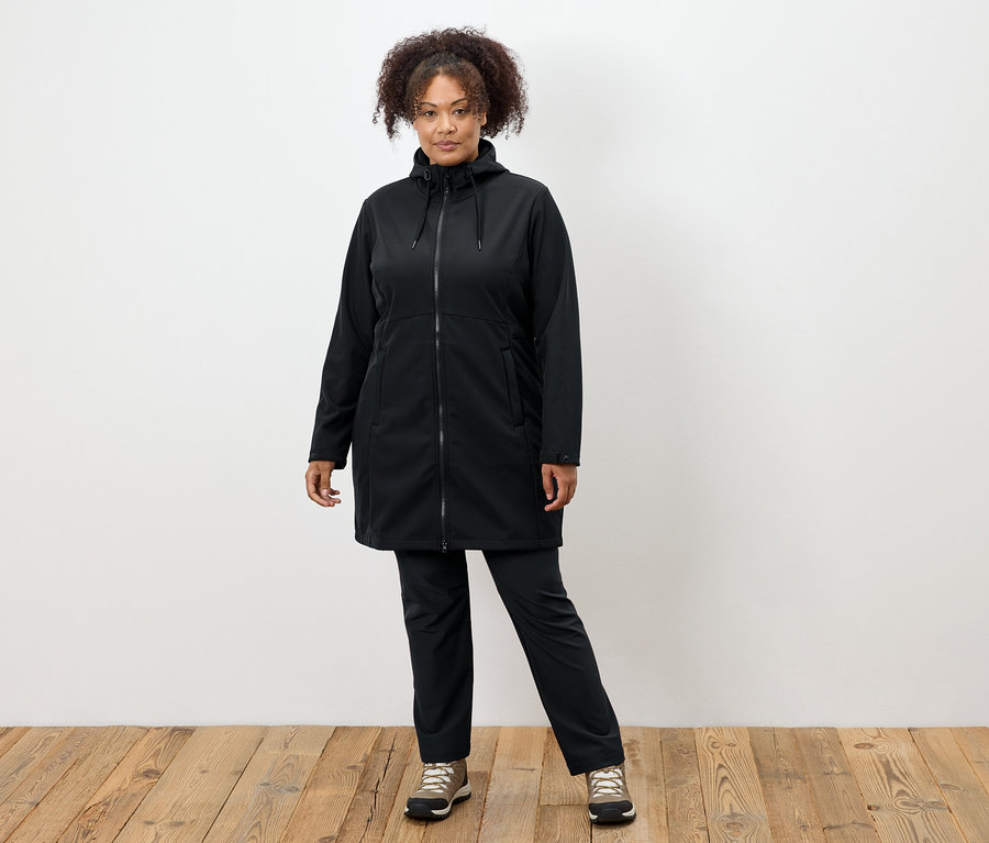 Femme en manteau et pantalon noirs debout sur un plancher en bois.