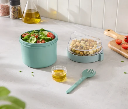 Salade composée dans un récipient turquoise avec des croûtons dans un récipient avec un couvercle transparent et de la vinaigrette et une fourchette à côté.