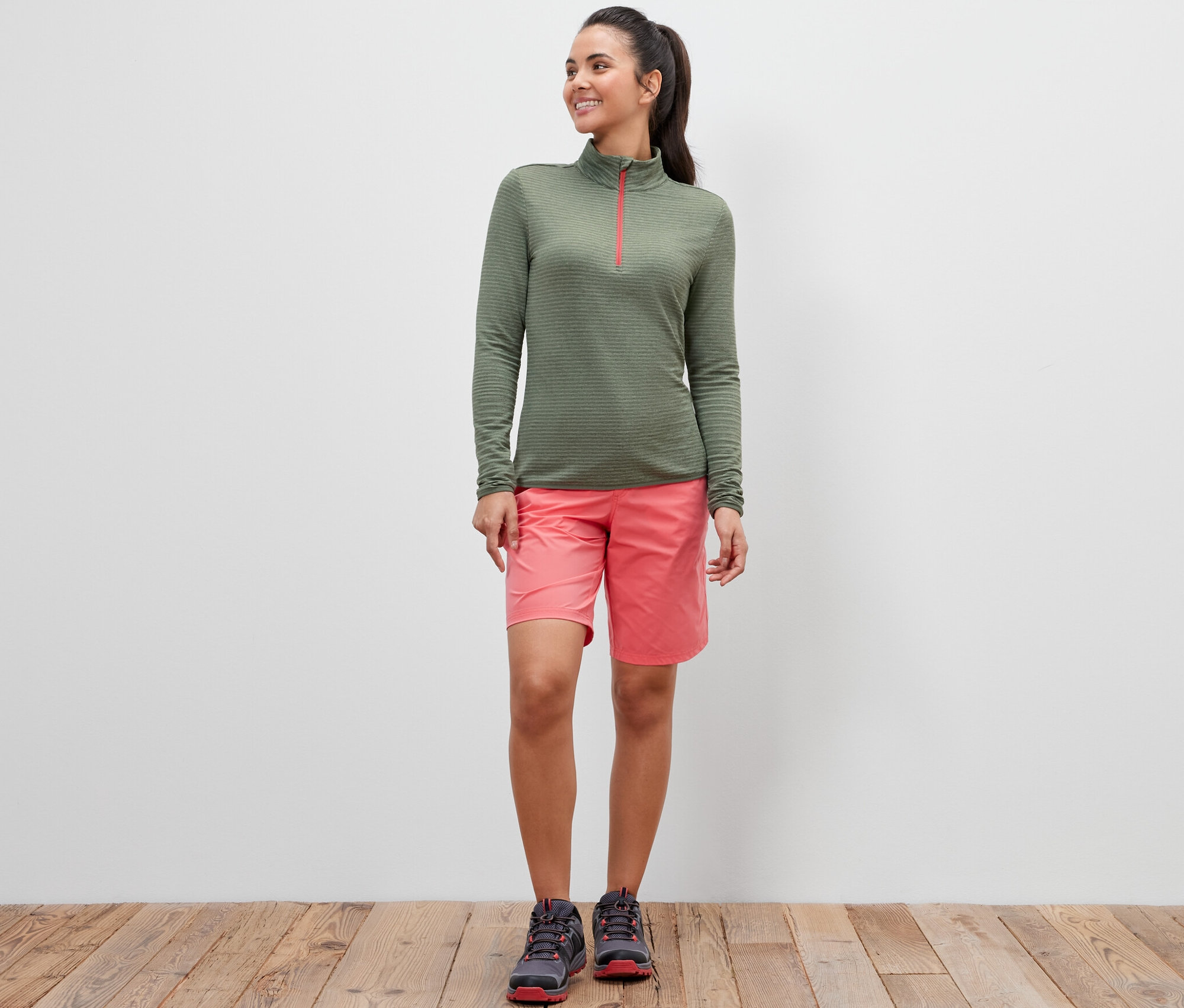 Une femme debout porte une chemise fonctionnelle outdoor verte, un short rose et des chaussures.