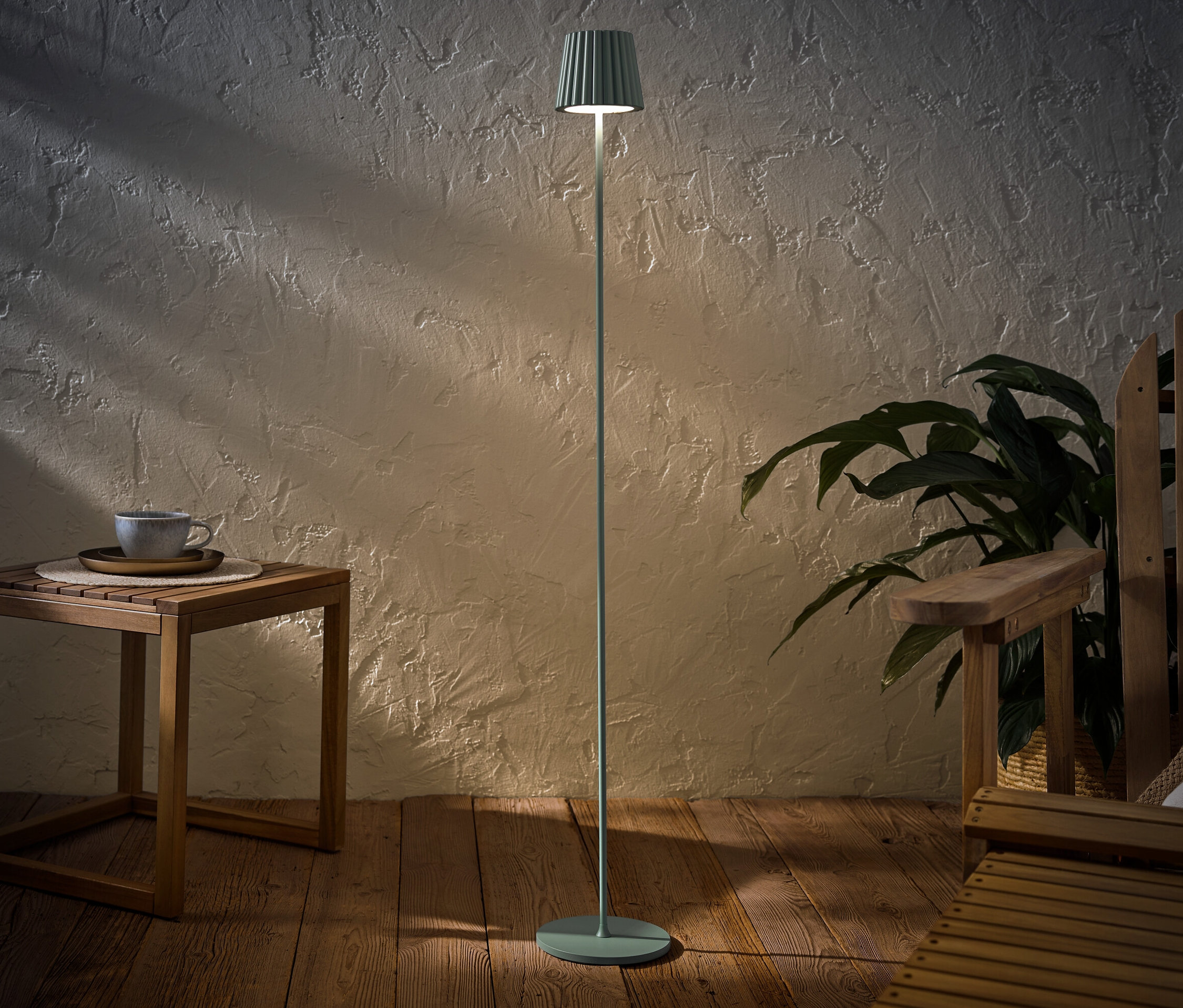 Un lampadaire d’extérieur à LED rechargeable, couleur sauge, est posé à côté d’une table en bois avec une tasse et d’une chaise en bois.