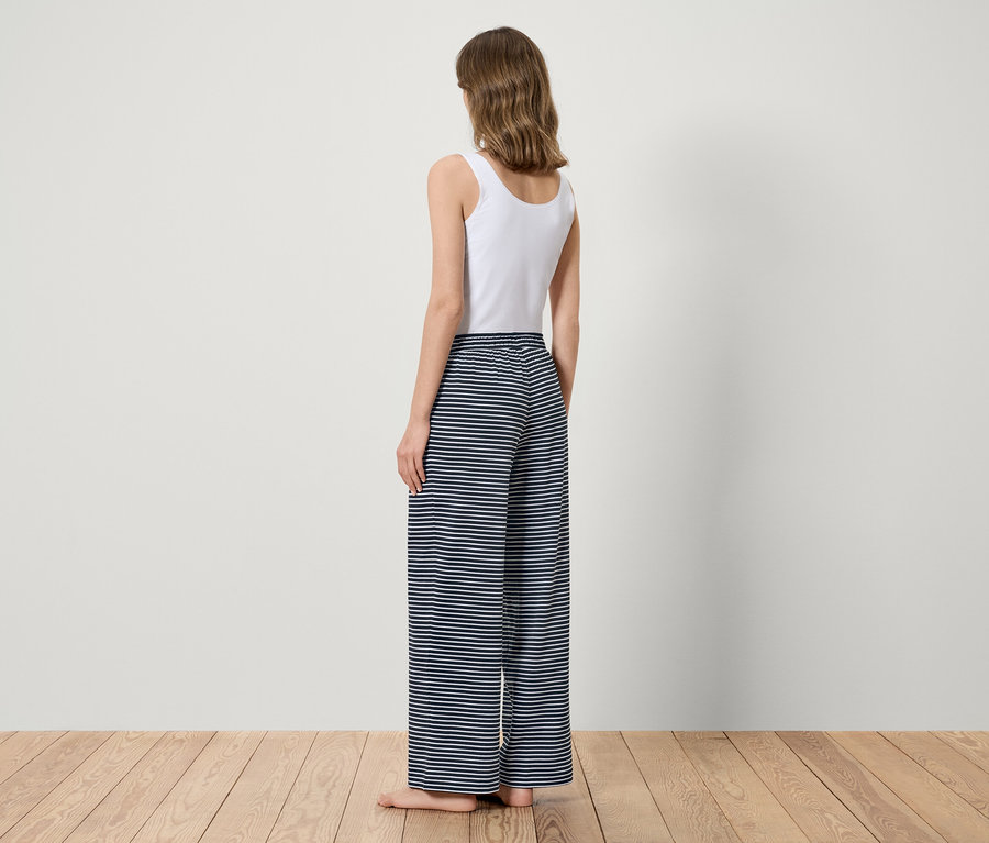 Une femme se tient dos à la caméra, vêtue d'un haut blanc et d'un pantalon à rayures bleues.