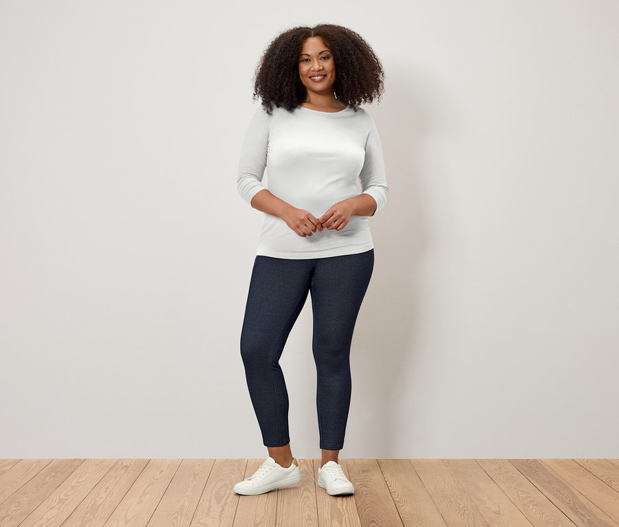 Une femme aux cheveux bouclés se tient sur un plancher en bois, portant un t-shirt blanc à manches trois-quarts et un legging bleu foncé.