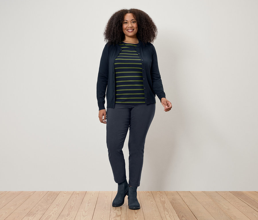 Une femme aux cheveux bouclés se tient sur un plancher en bois devant un mur blanc. Elle porte un cardigan bleu marine, un t-shirt rayé, un pantalon bleu marine et des bottes.