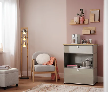 Salon avec fauteuil, lampe et commode avec machine à café. Sur la commode, un panier à capsules, blanc.