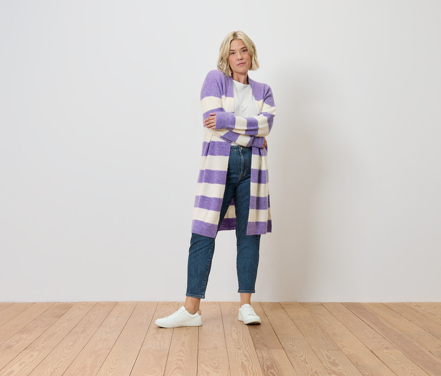 Une femme blonde se tient debout, les bras croisés. Elle porte un Mom Jeans – Coupe « Nyla », bleu moyen et un cardigan rayé violet et blanc.