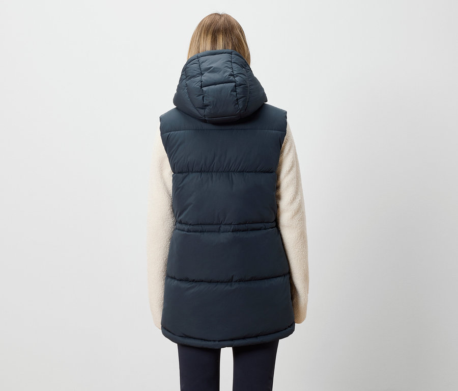 Vue arrière d'une femme aux cheveux blonds portant un gilet matelassé bleu marine à capuche et un pull crème.