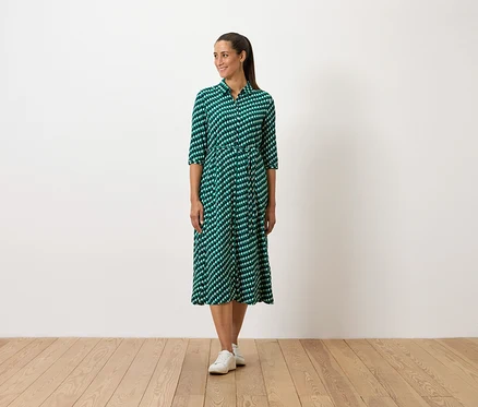Une femme debout porte une robe verte à motifs et des baskets blanches.
