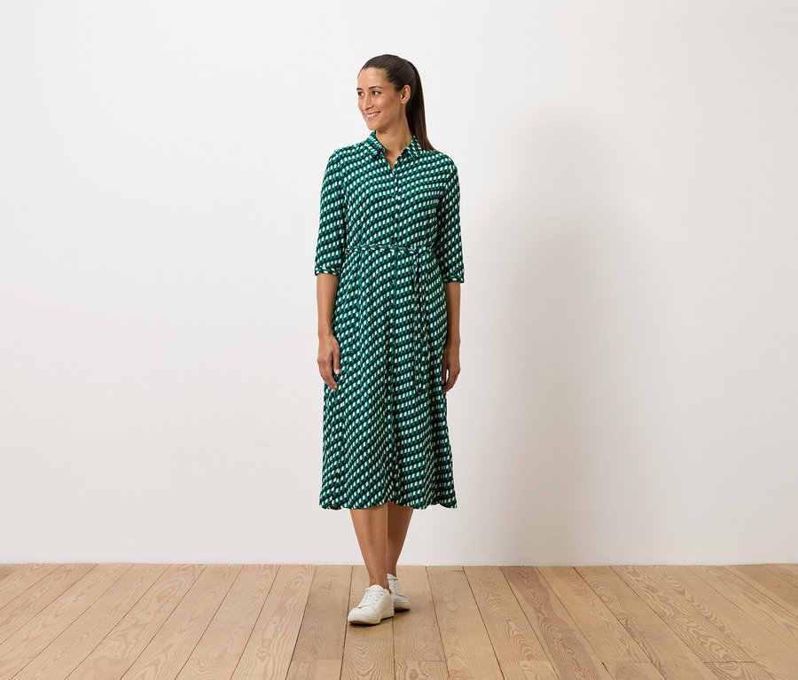 Une femme debout porte une robe verte à motifs et des baskets blanches.