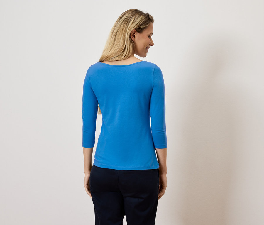 Une femme regarde par-dessus son épaule, portant un haut bleu à manches trois-quarts et un pantalon foncé.