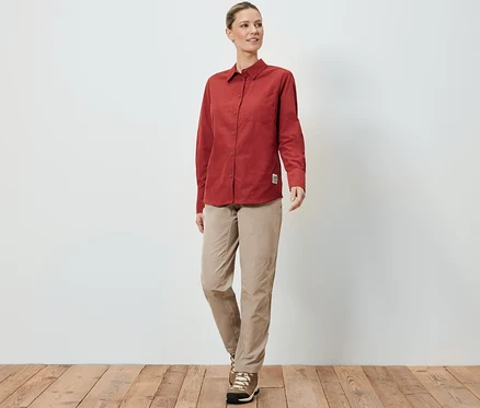Une femme portant une blouse d’extérieur avec protection anti-moustiques, un pantalon beige et des chaussures de randonnée, se tenant sur un plancher en bois devant un mur blanc.