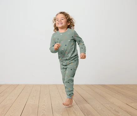 Une fillette aux cheveux bouclés, vêtue d'un pyjama vert imprimé de cœurs, court sur un plancher en bois.