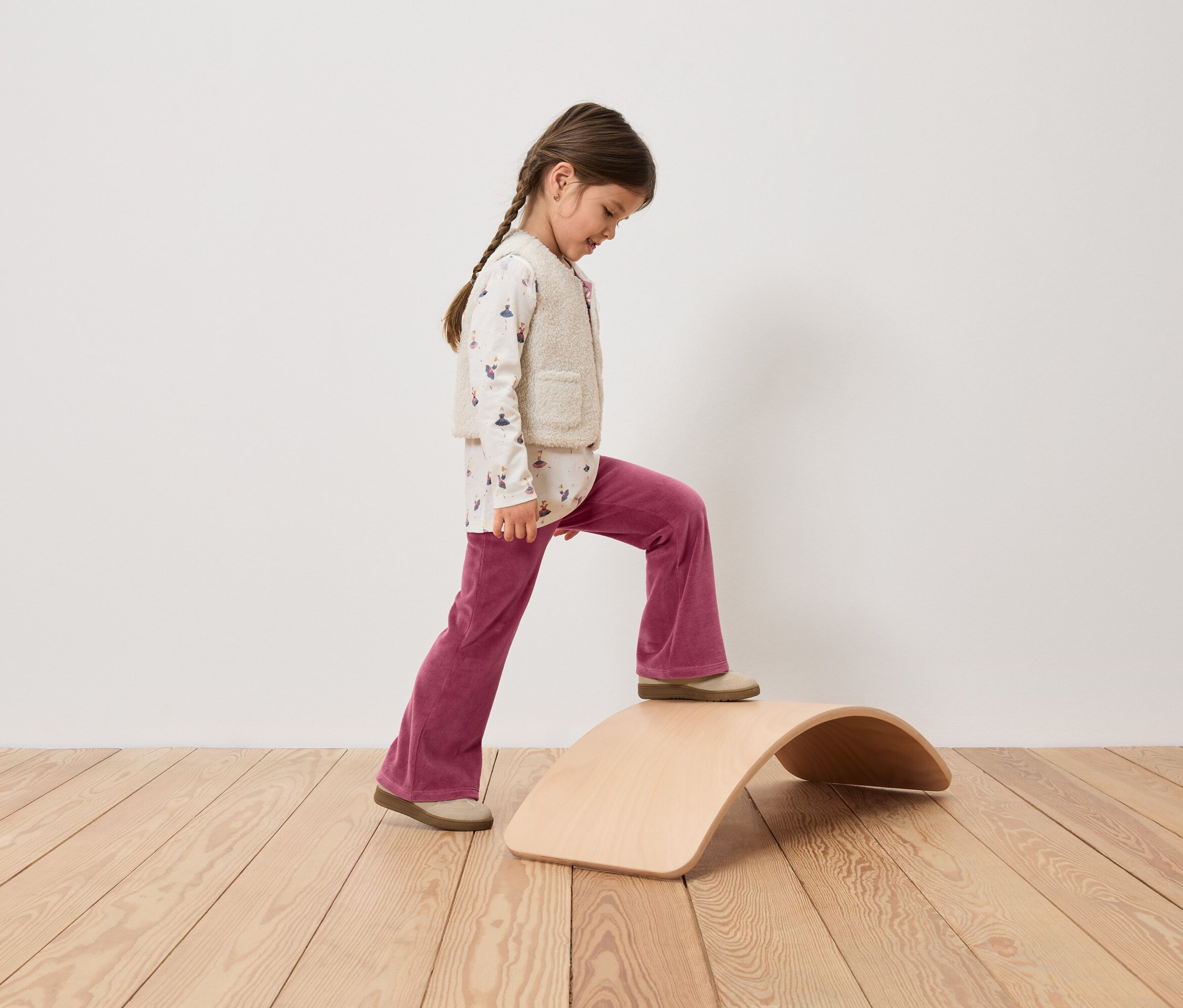 Une fille avec une tresse monte sur une planche d'équilibre en bois, vêtue d'un pantalon en velours rose, d'un haut imprimé et d'un gilet.