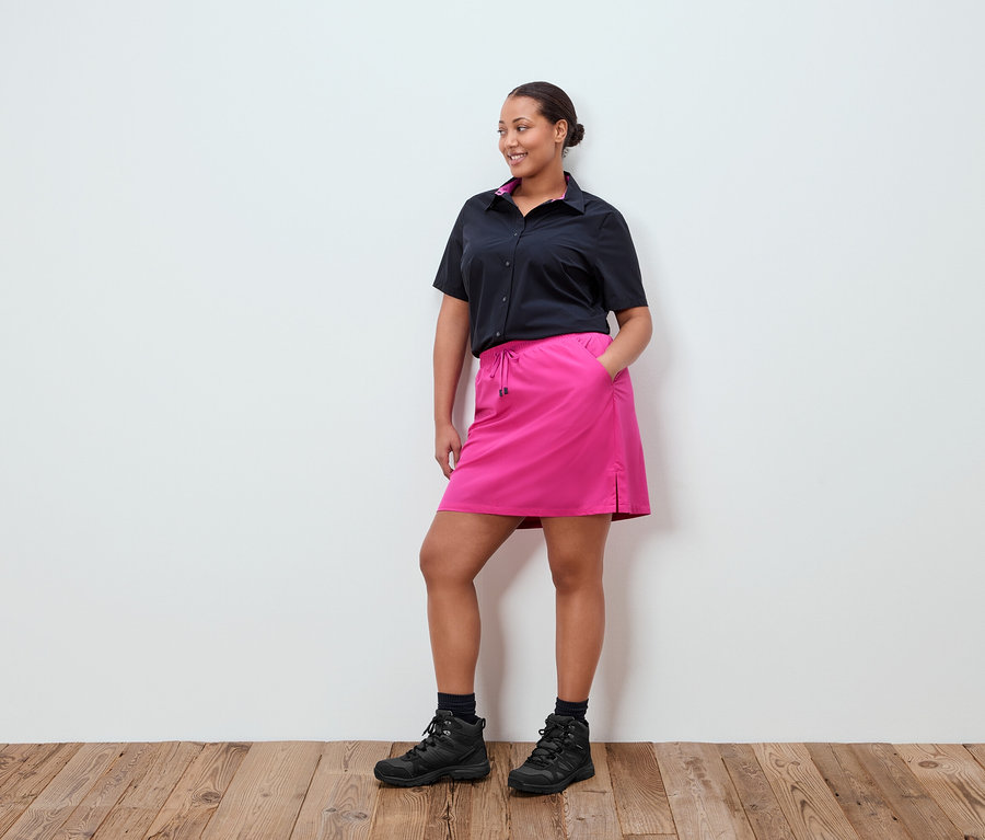 Une femme se tient devant un mur blanc, elle porte une chemise noire, une jupe rose et des chaussures noires.