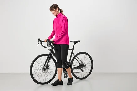 Femme portant une veste rose, un corsaire de cycliste noir et des chaussures noires, debout à côté d'un vélo noir.