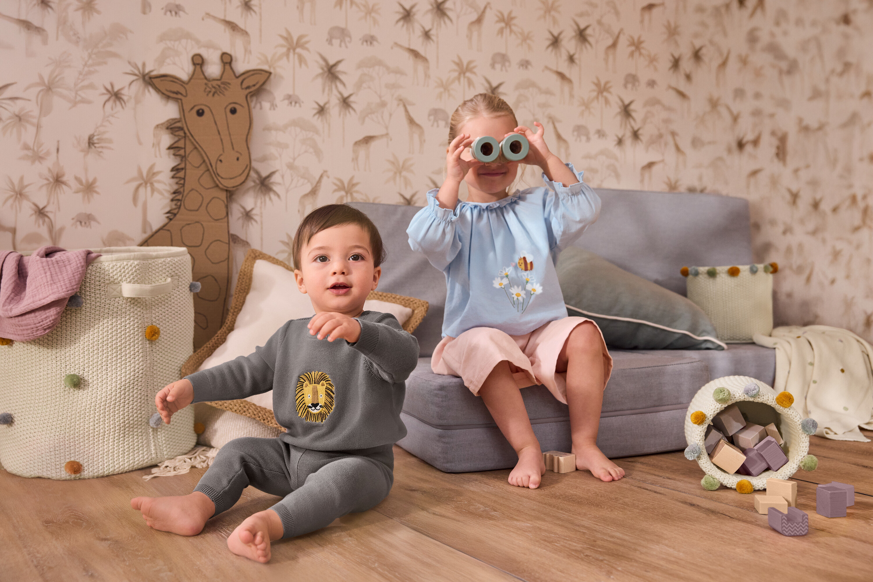 Un jeune enfant est assis à côté d'un grand panier de rangement pour chambre d'enfant. Une fille est assise sur un canapé gris et tient une couverture douillette en tricot pour bébé devant son visage. Elle porte un chemisier en chambray pour enfant et une culotte en velours côtelé pour enfant.