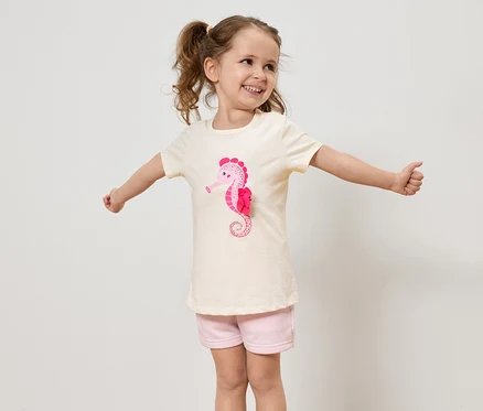 Une petite fille avec des tresses porte un t-shirt clair avec un hippocampe rose et un short rose, et elle tend les bras.
