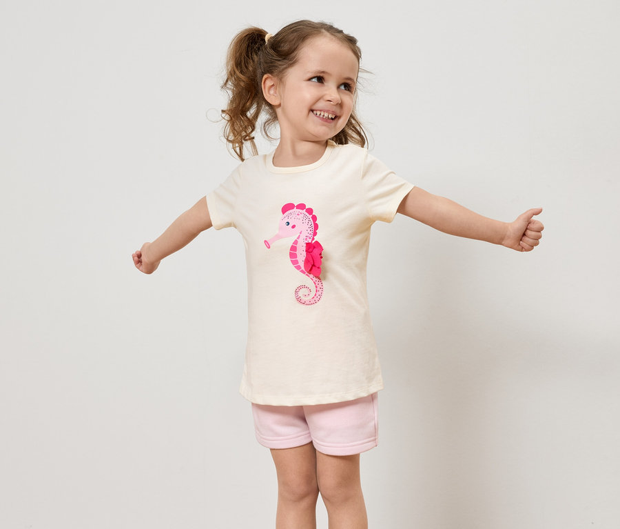 Une petite fille avec des tresses porte un t-shirt clair avec un hippocampe rose et un short rose, et elle tend les bras.