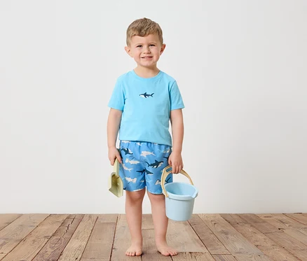 Un petit garçon blond se tient pieds nus sur un plancher en bois. Il porte un t-shirt bleu clair avec un requin et un short de bain bleu avec un motif de requin. Dans sa main gauche, il tient une pelle verte et dans sa main droite un seau bleu clair.