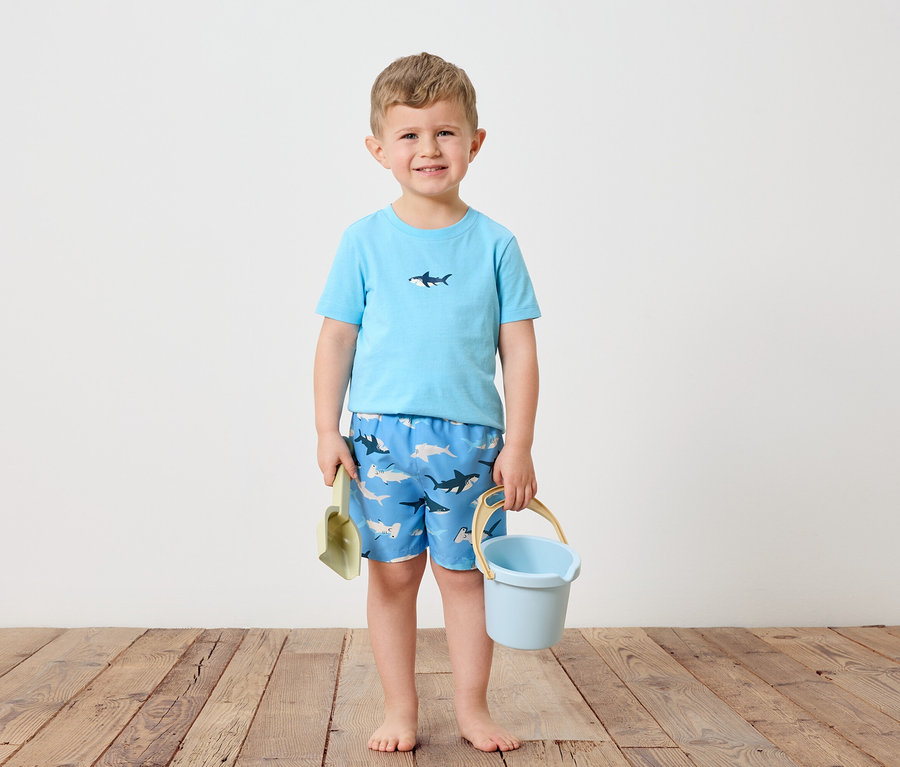 Un petit garçon blond se tient pieds nus sur un plancher en bois. Il porte un t-shirt bleu clair avec un requin et un short de bain bleu avec un motif de requin. Dans sa main gauche, il tient une pelle verte et dans sa main droite un seau bleu clair.