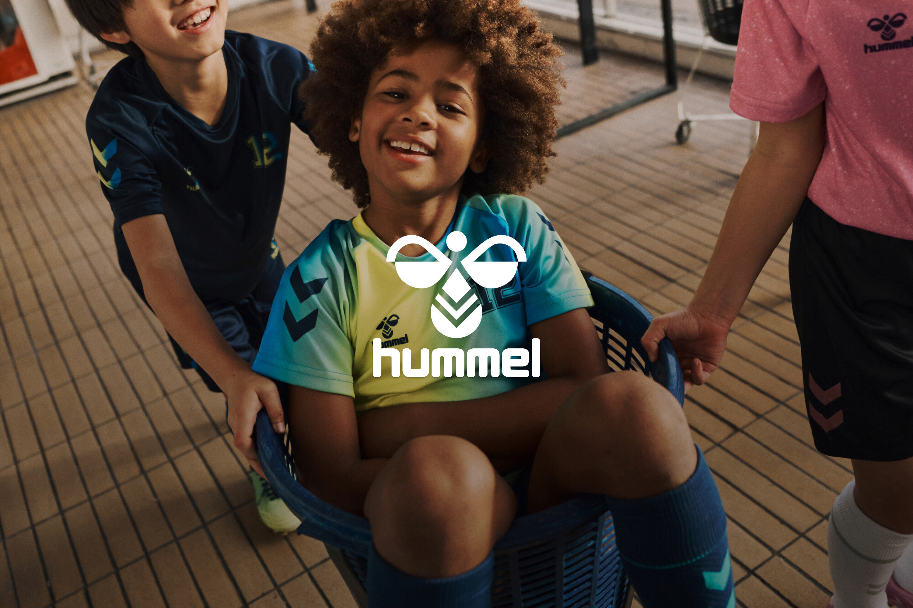 Trois enfants, dont un assis dans un panier, portent des vêtements de sport Hummel et sourient.