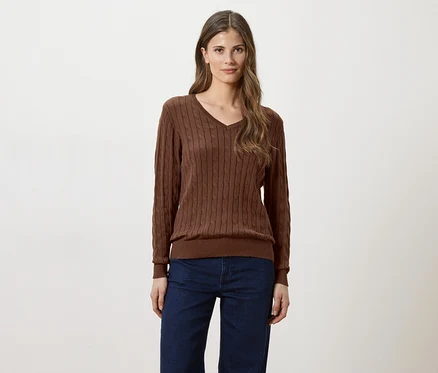 Une femme aux longs cheveux bruns portant un pull marron à col en V et un jean sur fond blanc.
