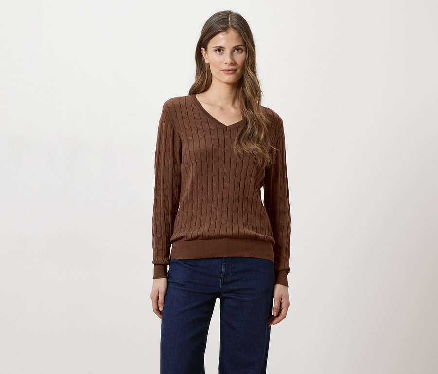 Une femme aux longs cheveux bruns portant un pull marron à col en V et un jean sur fond blanc.