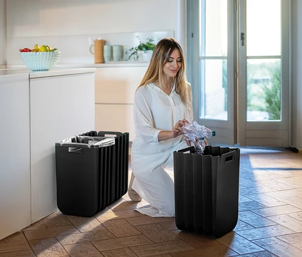 Une femme agenouillée jette une bouteille en plastique dans l'une des deux poubelles noires dans la pièce.