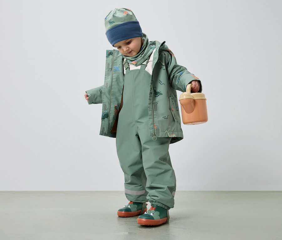 Un enfant portant des vêtements de pluie verts à motif de dinosaure tient un seau orange.