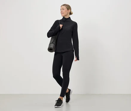 Femme portant un haut de sport noir, des leggings et des baskets avec un tapis de yoga sur l'épaule.