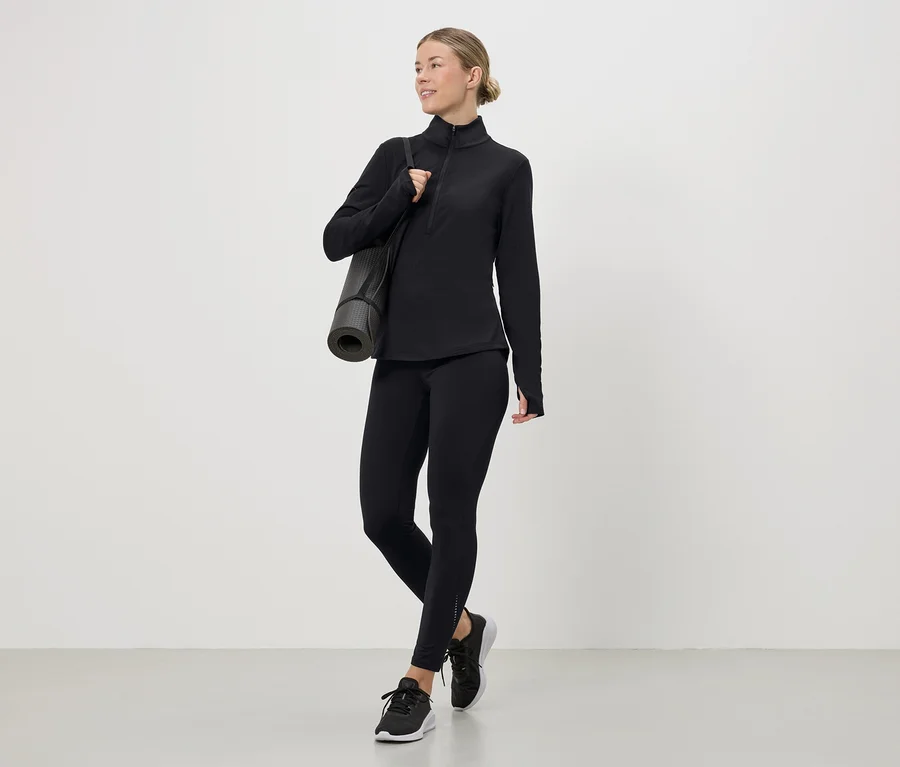 Femme portant un haut de sport noir, des leggings et des baskets avec un tapis de yoga sur l'épaule.