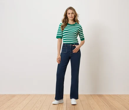 Une femme se tient debout, la main dans la poche de son pantalon, devant un mur blanc. Elle porte un haut rayé vert et blanc, un jean foncé et des chaussures blanches.