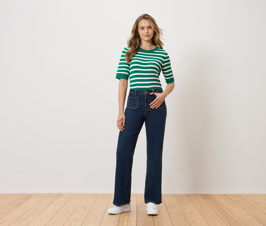 Une femme se tient debout, la main dans la poche de son pantalon, devant un mur blanc. Elle porte un haut rayé vert et blanc, un jean foncé et des chaussures blanches.