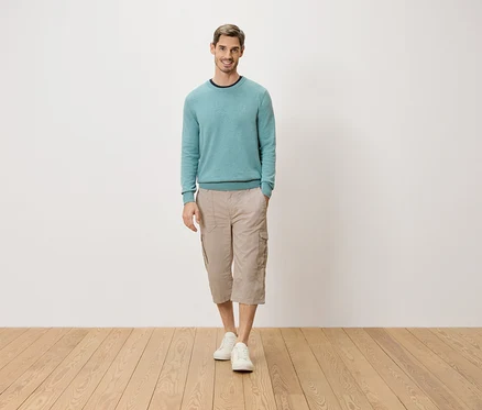 Un homme se tient debout, une main dans la poche, devant un mur blanc. Il porte un pull bleu clair, un pantacourt cargo beige et des chaussures blanches.