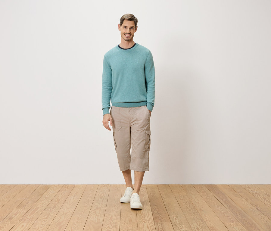 Un homme se tient debout, une main dans la poche, devant un mur blanc. Il porte un pull bleu clair, un pantacourt cargo beige et des chaussures blanches.