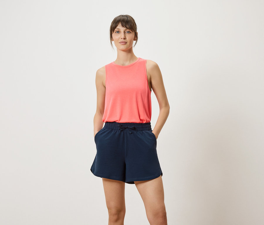Femme portant un débardeur corail et un short bleu marine avec les mains dans les poches.