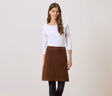 Femme portant une jupe en velours marron, des collants noirs et un haut blanc à manches longues sur fond clair.