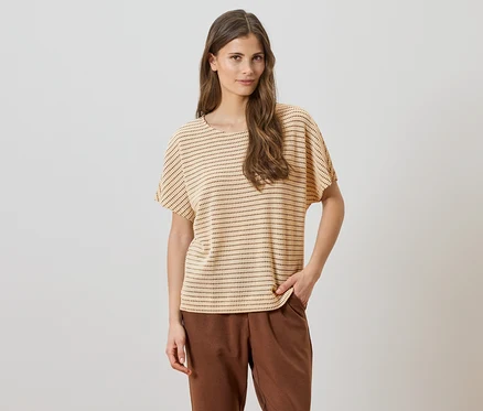 Une femme aux cheveux bruns ondulés porte un haut clair à manches courtes avec des points foncés et un pantalon marron sur un fond blanc.
