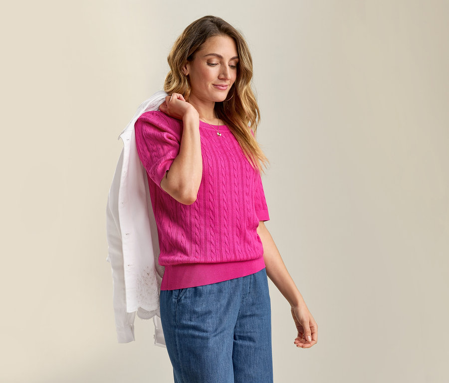 Femme portant une veste en jean avec broderie ajourée sur l'épaule et un pull en maille fine avec torsades rose avec un jean.