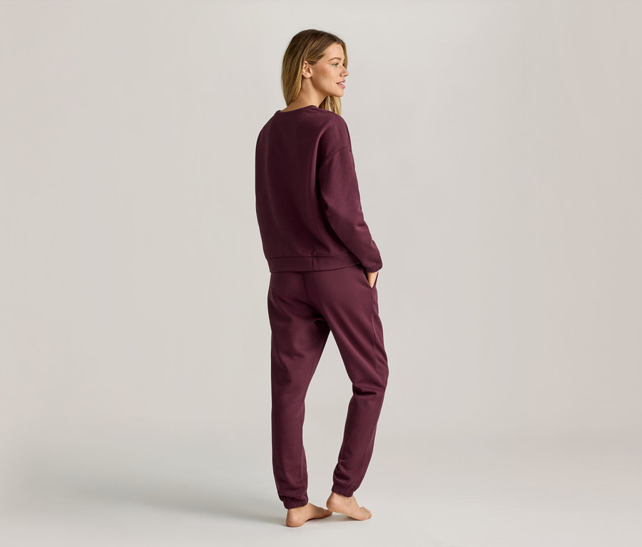 Une femme aux cheveux blonds porte un sweat-shirt bordeaux et un pantalon de jogging, vue de dos.