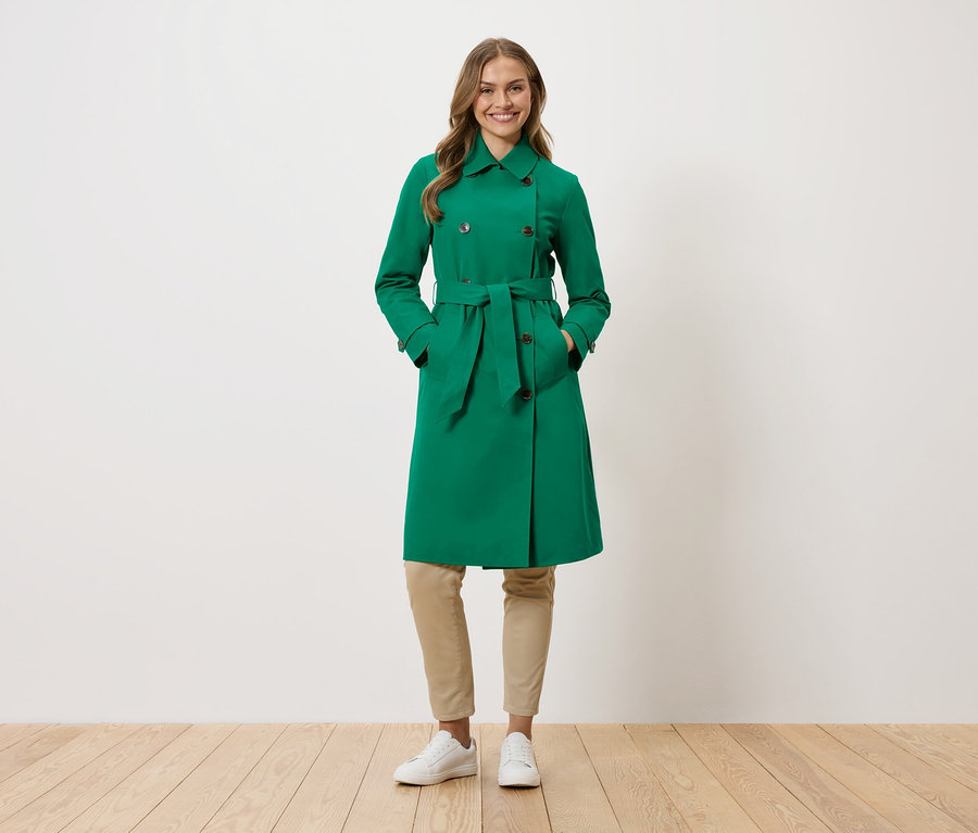 Une femme se tient debout, les mains dans les poches d'un trench-coat vert.