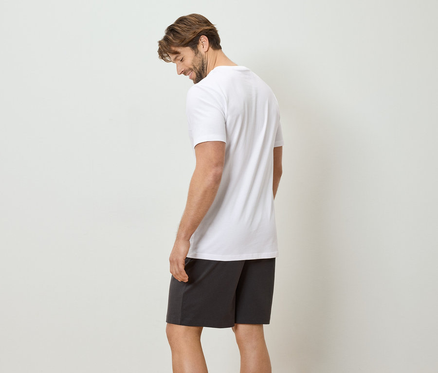 Un homme portant un t-shirt blanc et un short foncé se tient devant un mur blanc.